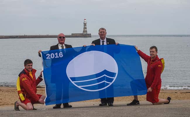 Councillor John Kelly, (3rd from left) Portfolio Holder for Public Health, Wellness and Culture, Sunderland City Council, with l-r Alexander Robinson lifeguard, Tom Parkin chairman Sunderland Seafront Traders Association and Phil Johnson lifeguard with the Blue Flag for Roker and Seaburn beaches.